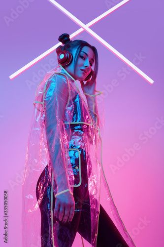 Beautiful asian woman in a raincoat listens music in headphones around colourful neon