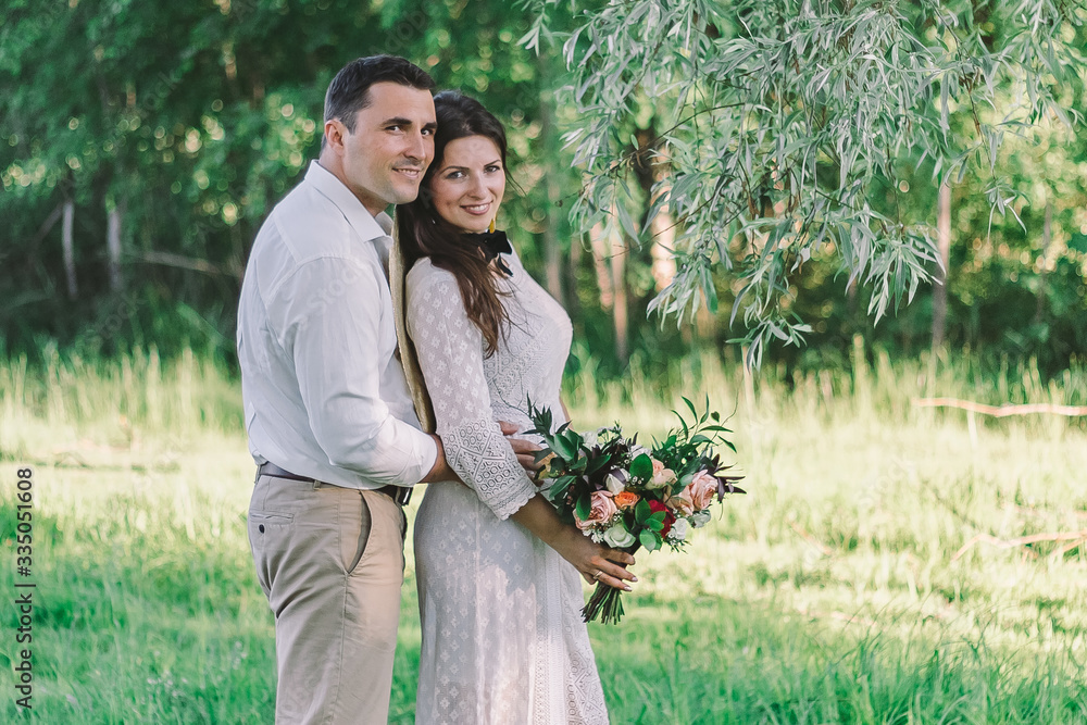 Wedding. The groom in a white shirt and light pants, the bride in a beautiful dress and a straw hat with a beautiful wedding bouquet in her hands. Hug each other look at a tree. Wedding. The groom in a white shirt and light pants, the bride in a beautiful dress and a straw hat with a beautiful wedding bouquet in her hands. Hug each other look at a tree.