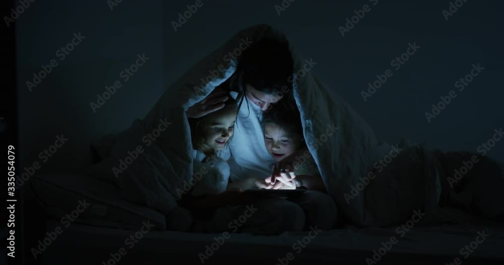 An young happy mother and her daughters are using a tablet in the dark under the blanket. Concept of family, entertainment, education, technology, motherhood, togetherness An young happy mother and her daughters are using a tablet in the dark under the blanket. Concept of family, entertainment, education, technology, motherhood, togetherness