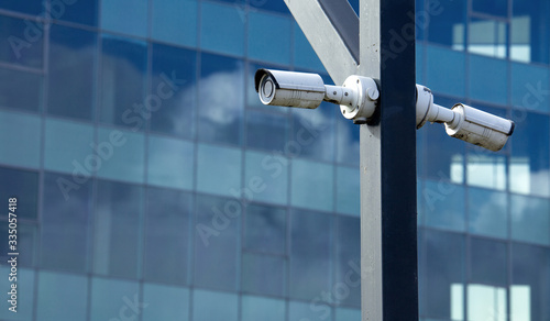 Two surveillance cameras mounted on a pillar in the background of a glass building