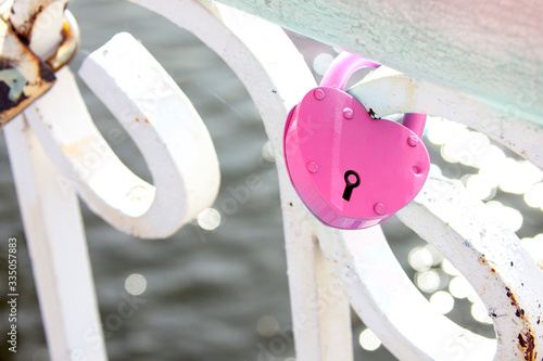 фотографія A pink heart-shaped padlock hanging on a white fence against a water background