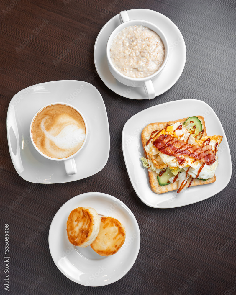 Breakfast consists of oatmeal porridge with cheesecakes and cappuccino with sandwich, top view. Breakfast consists of oatmeal porridge with cheesecakes and cappuccino with sandwich, top view.