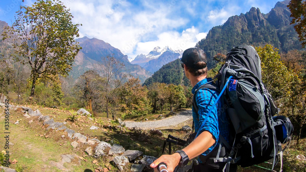A man with a big black backpack hikes along a pathway on Annapurna Circuit Trek in Himalayas, Nepal, while taking a selfie. There are massive mountain chains around him. Exploration and adventure