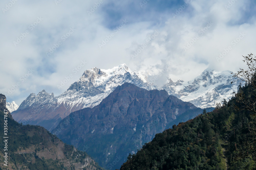 View on Himalayas, Annapurna Circuit Trek, Nepal. The view is disturbed by dense tree crowns in the front. High, snow caped mountains peaks catching the sunbeams. Serenity and calmness. Barren slopes