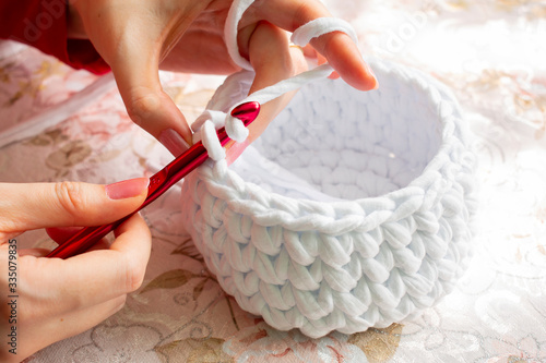 Young woman while crocheting on a patterned tablecloth, close up. Stay at home leisure activity idea. Basket made of white T-shirt yarn, with red crochet needle. Pale pink nails. photo