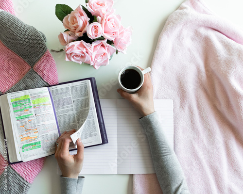Devotional Bible study. Woman's hand is turning a page of the Bible. With pink roses, black tea and coffee. Background pink, white and grey colors.