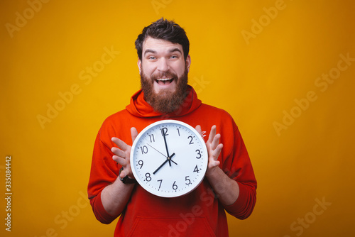 Oh no, what time is it. Bearded surprised man is holding a white clock on yellow background.