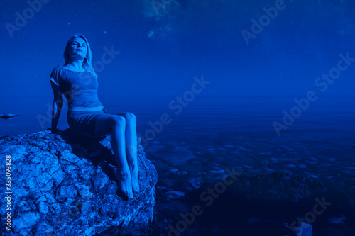Young relaxed caucasian woman sit on a stone and enjoys summer night on the background of the starry sky during the long-awaited vacation on a summer night. Rest and recovery concept