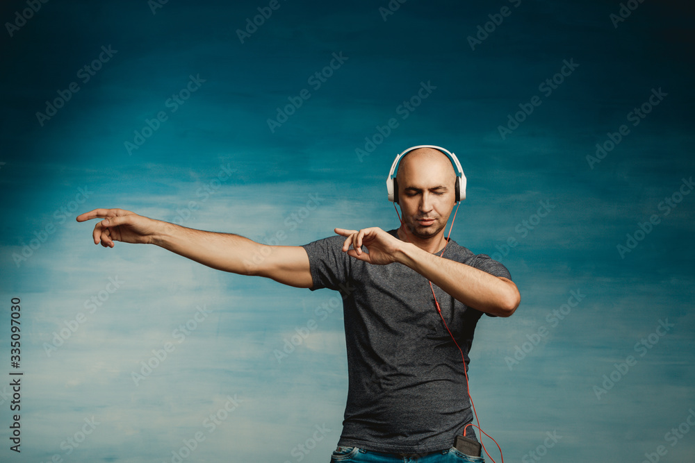 Cool bald man in a gray T-shirt and jeans jumping and fooling around and dancing in headphones on a blue background. Cool bald man in a gray T-shirt and jeans jumping and fooling around and dancing in headphones on a blue background.