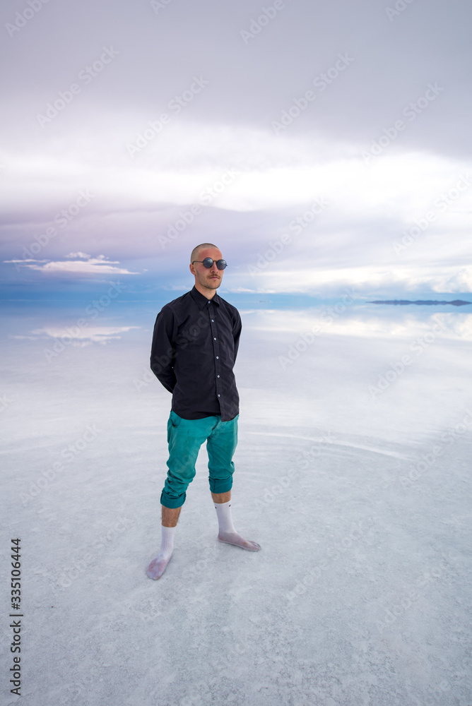 Sun Burnt Caucasian Bold Man with Sunglasses in Black Shirt and Green Pants Poses in Flooded Uyuni Salt Flats at Sunset Sun Burnt Caucasian Bold Man with Sunglasses in Black Shirt and Green Pants Poses in Flooded Uyuni Salt Flats at Sunset