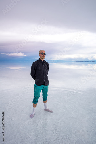 Sun Burnt Caucasian Bold Man with Sunglasses in Black Shirt and Green Pants Poses in Flooded Uyuni Salt Flats at Sunset