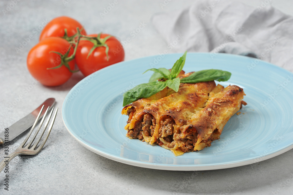 Cannelloni with minced meat on a blue plate. Cutlery and ripe tomatoes complete the composition. Light background. Close-up. Cannelloni with minced meat on a blue plate. Cutlery and ripe tomatoes complete the composition. Light background. Close-up.