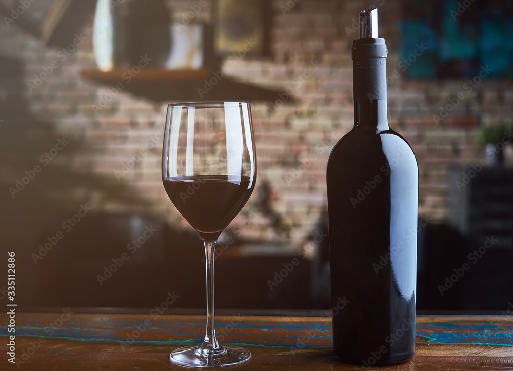 Glass of red wine and wine bottle on colorful wooden table at home, brick wall in the background.