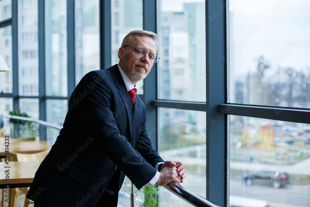 Adult male businessman, teacher, mentor looks out the large window of his office. Work in the office. Development of a new business project Adult male businessman, teacher, mentor looks out the large window of his office. Work in the office. Development of a new business project