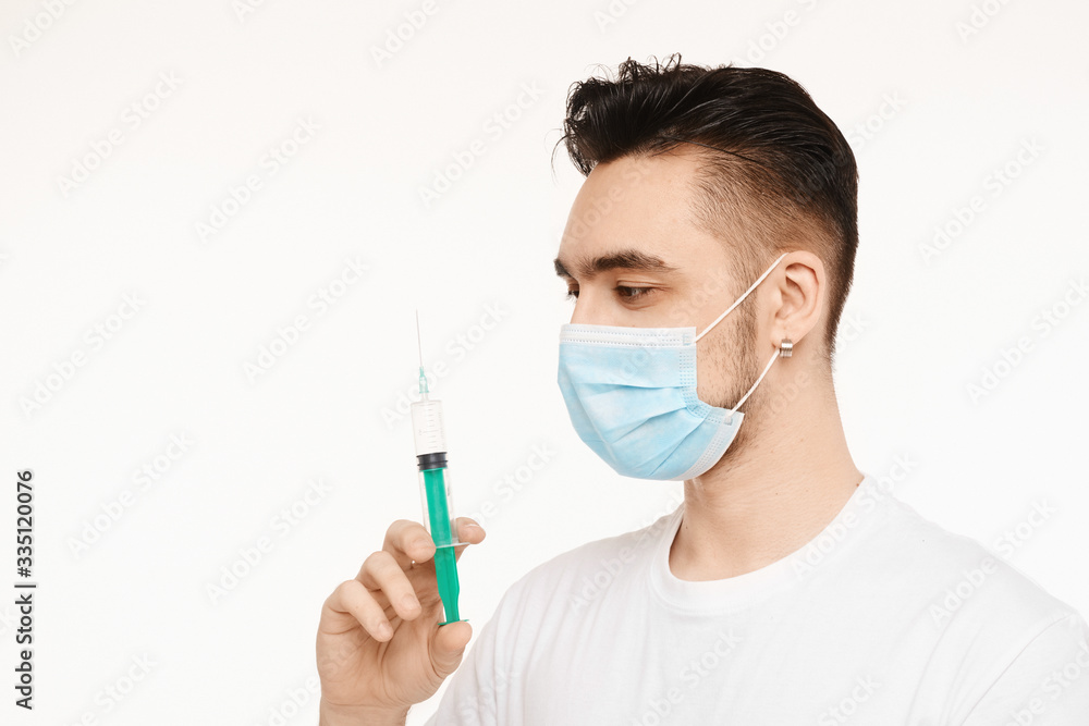 Handsome men in a white t-shirt and protective mask holding a syringe and looking on the needle, isolated in a white background. Medical advertisement mockup with copy space Handsome men in a white t-shirt and protective mask holding a syringe and looking on the needle, isolated in a white background. Medical advertisement mockup with copy space