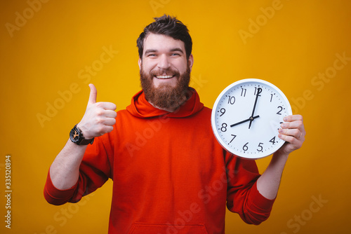 Track your time, it flies. Photo of a happy man showing thumb up and holding a white clock on yellow background.
