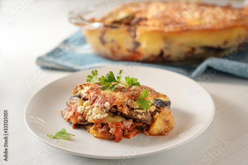 Moussaka, traditional Greek baked casserole of eggplants, potatoes, minced meat and tomatoes served with parsley garnish on a plate on a white table