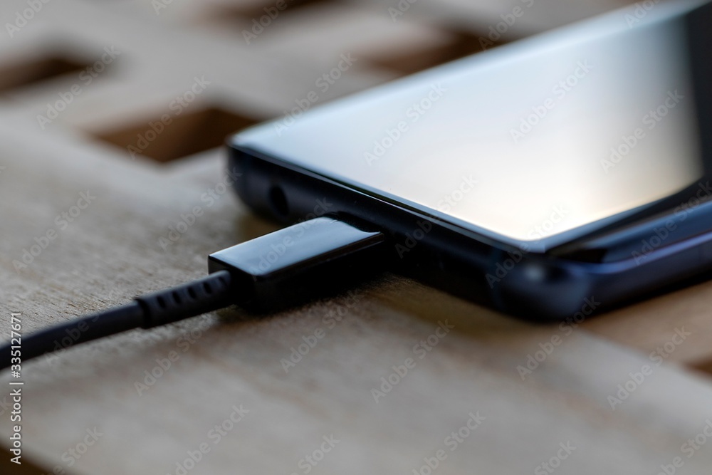 A close up portrait of a phone lying on a wooden table getting its battery charged using a USB cable connected to its port. The smartphone had a flat battery.