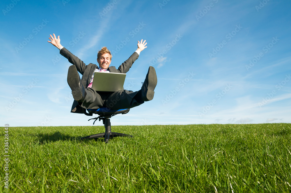Happy businessman celebrating online success sitting on an office chair outdoors with his laptop computer in the middle of a green meadow Happy businessman celebrating online success sitting on an office chair outdoors with his laptop computer in the middle of a green meadow