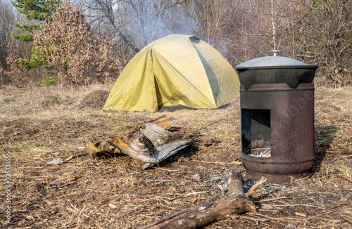 A tourist tent stands in a clearing and food is being prepared in front of it in a metal stove