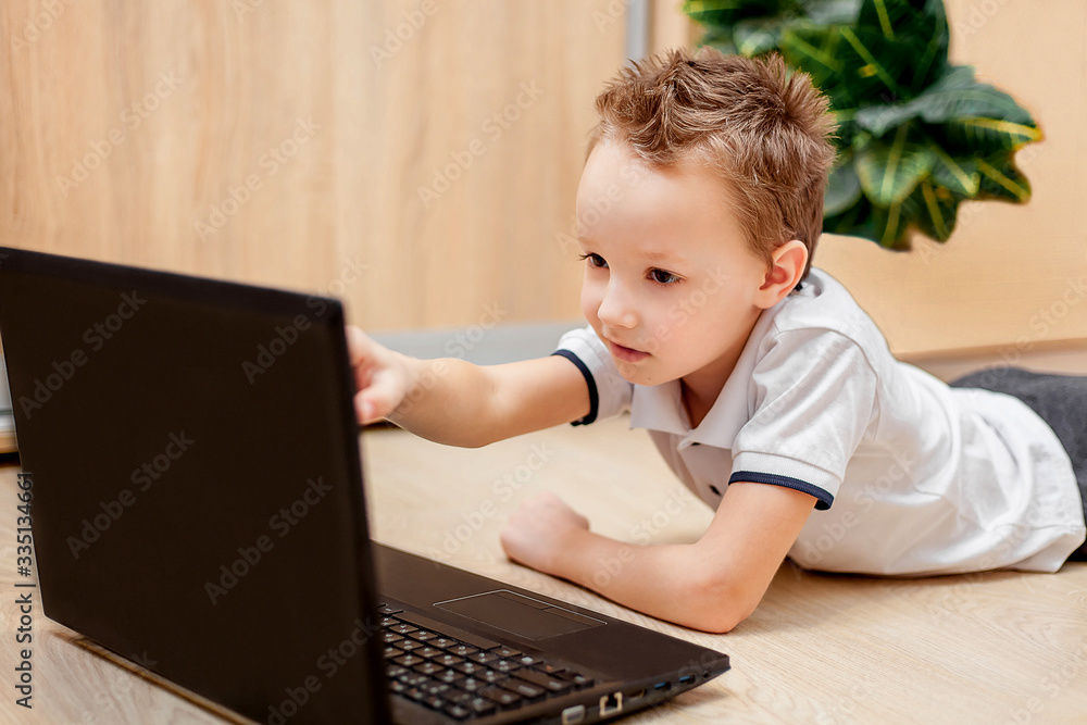 Remote online home training. The child lies on the floor and works behind the computer. The schoolboy looks at the computer and points with his hand to the monitor. Quarantine. Coronavirus. Remote online home training. The child lies on the floor and works behind the computer. The schoolboy looks at the computer and points with his hand to the monitor. Quarantine. Coronavirus.