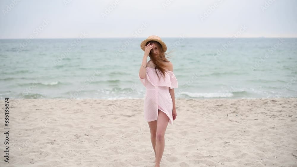 Beautiful girl with long brunette hair in straw hat and dress walks along sea Beautiful girl with long brunette hair in straw hat and dress walks along sea