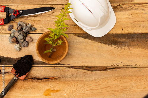 White engineering helmet, garden shears, flower pot, hand shovel with black soil and gravel on wooden background. Concept of engineering services, design and construction of houses and gardens