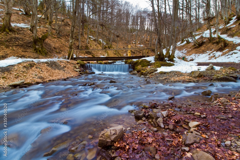 clearwater river Pylypets with fast flow and a little bridge in the forest, snow, fallen leaves and stones on banks of the creek, Carpathian Mountains in Transcarpathia, ecology, long exposure