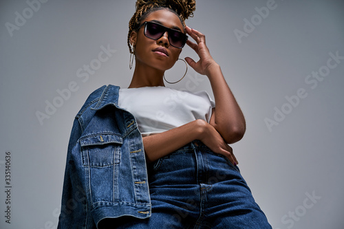 Confident lady in white t-shirt in jeans with denim jacket posing and looking at camera holds hands on glasses and hip. Fashion concept