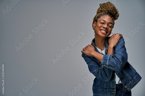 Happy smiling woman with earrings in denim clothing with braids in glasses hugs herself. Emotions concept