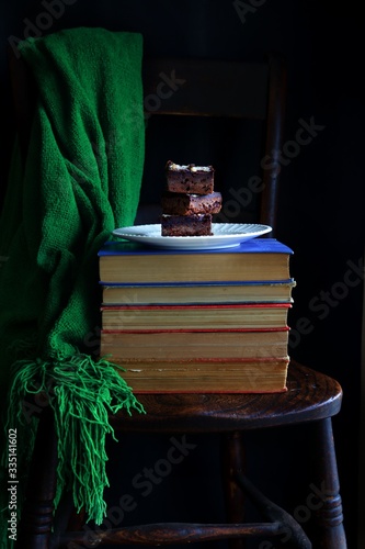 Banting chocolate brownies on a white plate on a pile of books on a wooden chair with a vivid green blanket hanging over the back of the chair against a dark background. photo