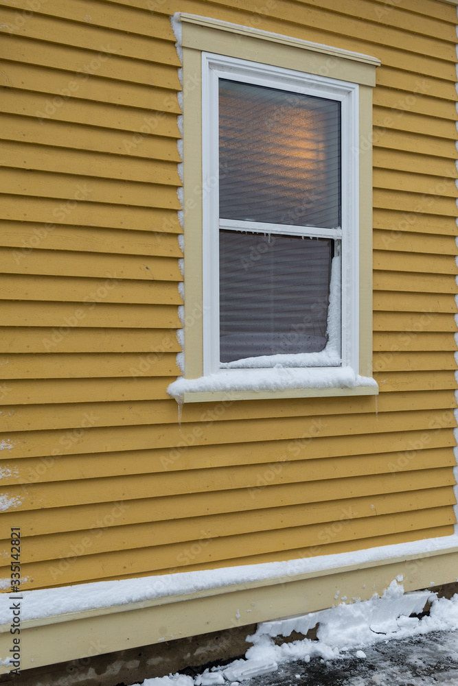 Fototapeta premium The exterior of a yellow coloured wall of narrow clapboard. In the center of the building, there's a double-hung window that is closed and has snow on the ledge and light at the top of the window. 