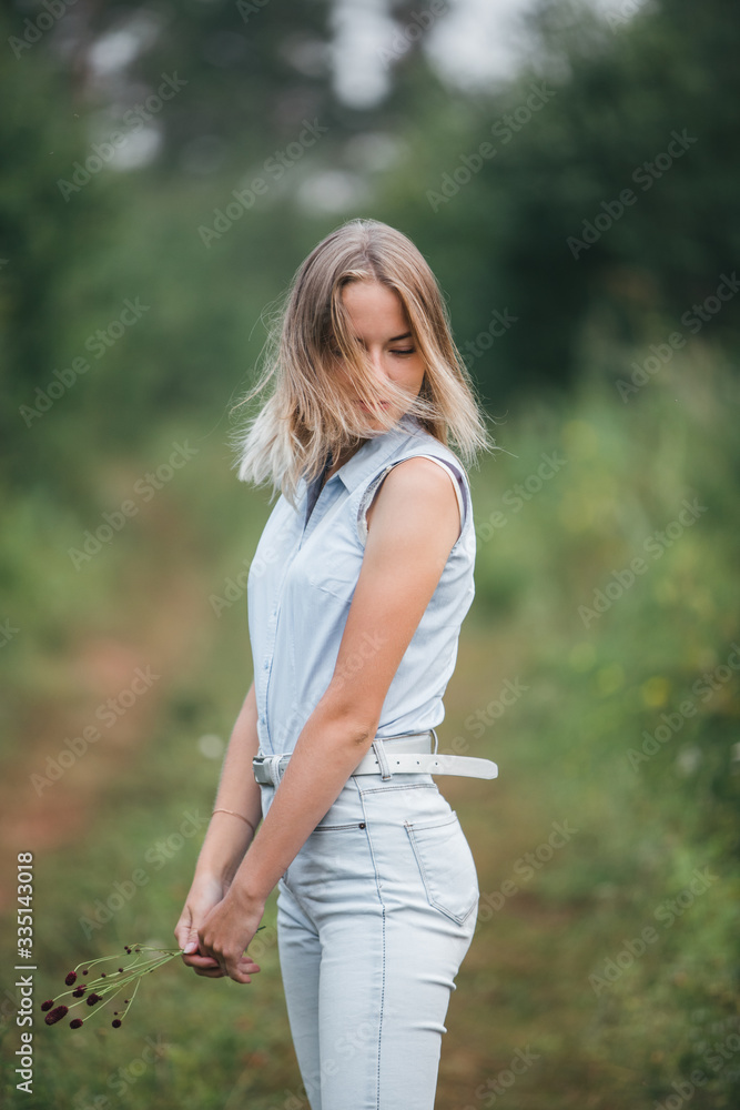A young blonde girl stands on a forest road with flowers in her hands and turns her head. Hair fluttering in the wind. A young blonde girl stands on a forest road with flowers in her hands and turns her head. Hair fluttering in the wind.