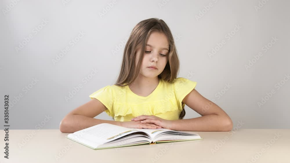 Girl flips through the pages of a book, and reads a book sitting at a table on a white background. The girl learns to read and runs her finger over the letters. Girl flips through the pages of a book, and reads a book sitting at a table on a white background. The girl learns to read and runs her finger over the letters.