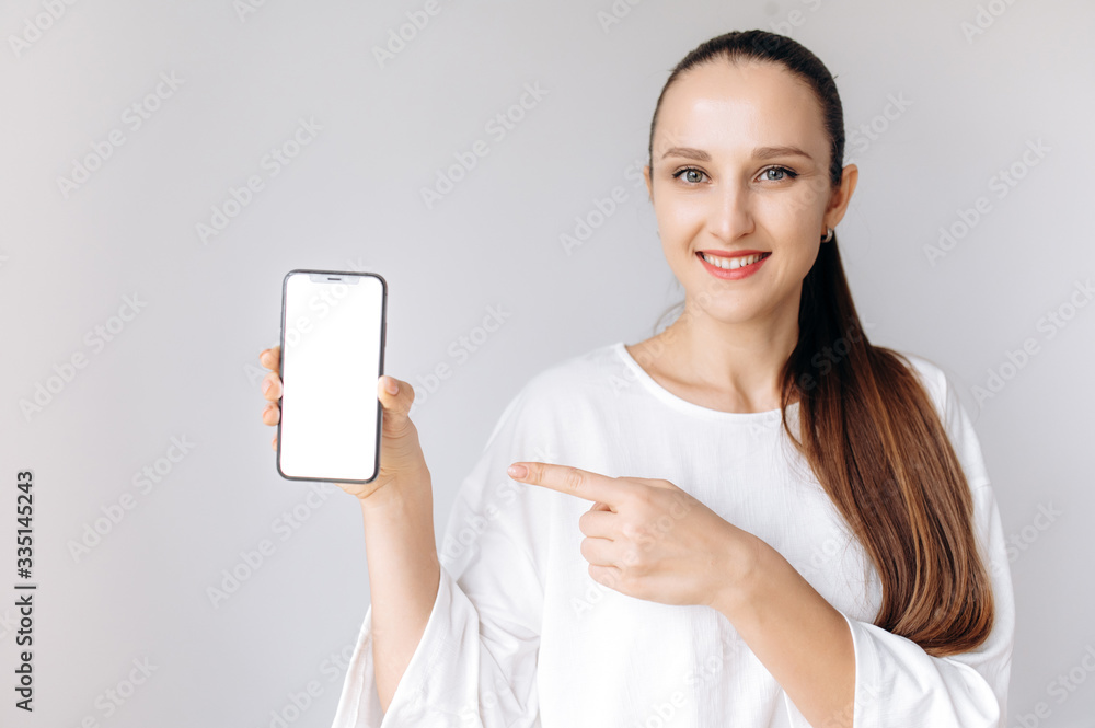 Attractive smiling girl holding a phone in her hand with a blank white screen standing on a white background Attractive smiling girl holding a phone in her hand with a blank white screen standing on a white background