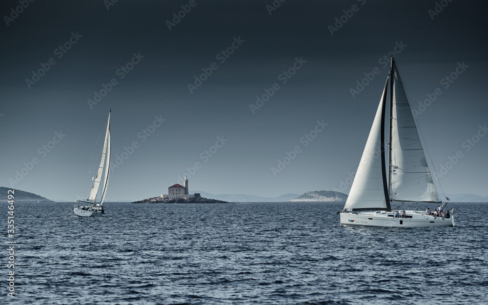 Sailboats compete in a sailing regatta at sunset, sailing race, reflection of sails on water, multi-colored spinaker, boat number aft boats, big white clouds, the island with a lighthouse