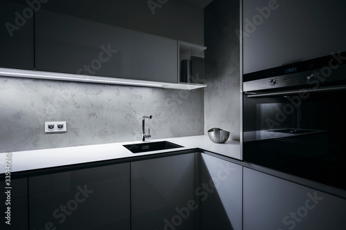 Modern white kitchen in minimalist design, in night, with light strip turned on, modern oven, granite sink, wall-sockets and premium materials such as glass, concrete, aluminum and stainless steel.