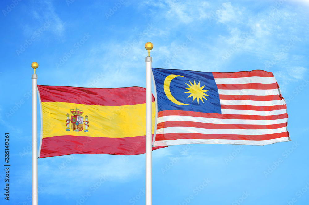 Spain and Malaysia two flags on flagpoles and blue cloudy sky Spain and Malaysia two flags on flagpoles and blue cloudy sky