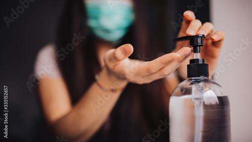 Young asian woman with long hair wearing medical mask and sanitizing her hands at home. Concept of coronavirus. Horizontal image, selectvie focus on hands and bottle of hand sanitizer gel.