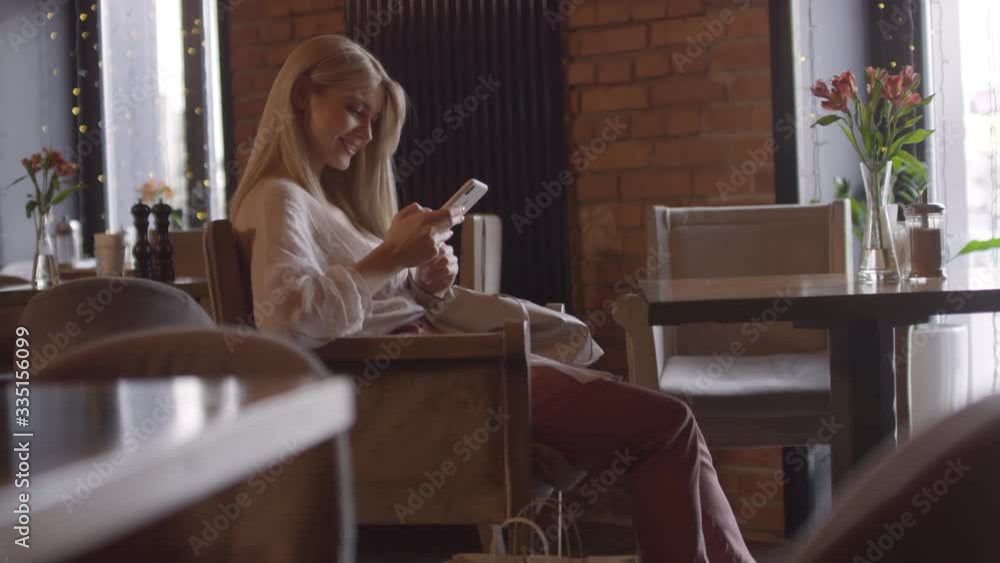 Handheld shot of happy woman with blond hair sitting at table in cafe and smiling while typing on mobile phone Handheld shot of happy woman with blond hair sitting at table in cafe and smiling while typing on mobile phone