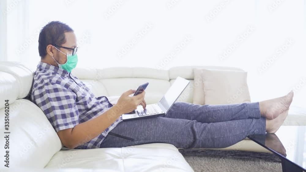 Man working with laptop and cellphone while wearing a mask on the sofa at home. Shot in 4k resolution Man working with laptop and cellphone while wearing a mask on the sofa at home. Shot in 4k resolution