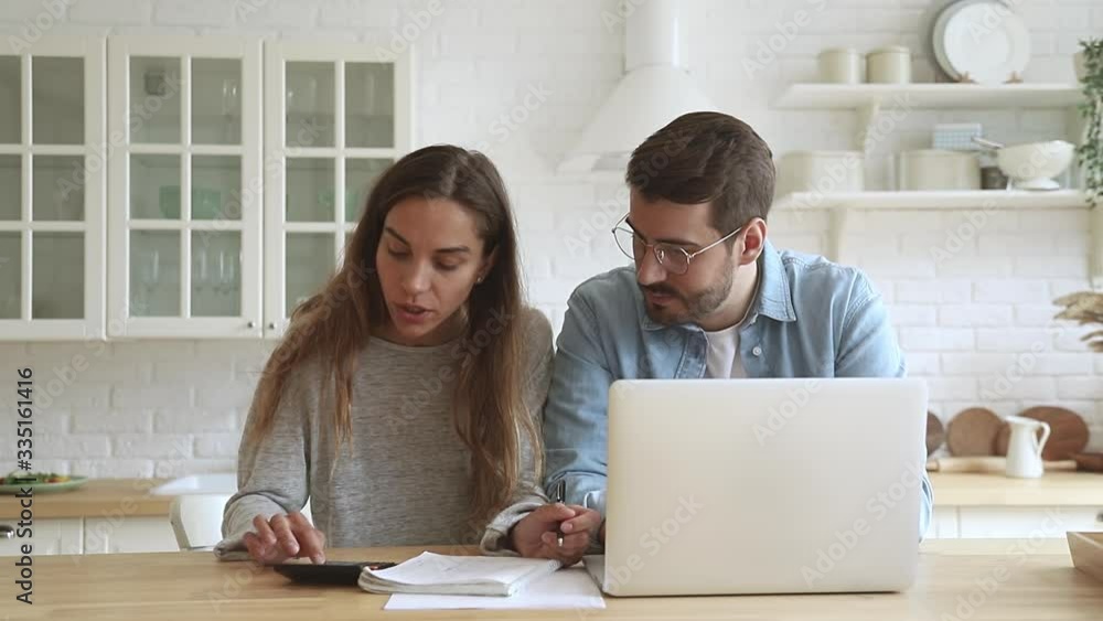 Concentrated millennial mixed race family couple managing monthly budget together in kitchen. Serious young woman discussing expenses, mortgage payments, taxes, utility bills with husband at home. Concentrated millennial mixed race family couple managing monthly budget together in kitchen. Serious young woman discussing expenses, mortgage payments, taxes, utility bills with husband at home.