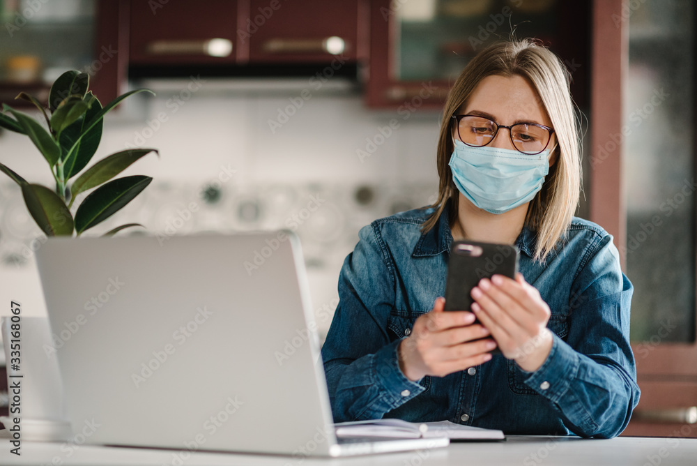 Coronavirus. Girl in mask checking social apps in phone. Woman work, learns and using laptop computer. Quarantine. Stay at home. Freelancer. Writing, typing. Communication and technology concept. Coronavirus. Girl in mask checking social apps in phone. Woman work, learns and using laptop computer. Quarantine. Stay at home. Freelancer. Writing, typing. Communication and technology concept.