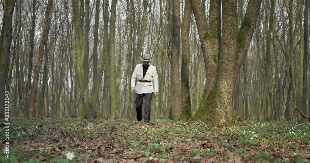 Full length portrait of bearded man in black trousers, white jacket and hat walking among green forest with walking stick. Mature botanist spending time on fresh air. Full length portrait of bearded man in black trousers, white jacket and hat walking among green forest with walking stick. Mature botanist spending time on fresh air.