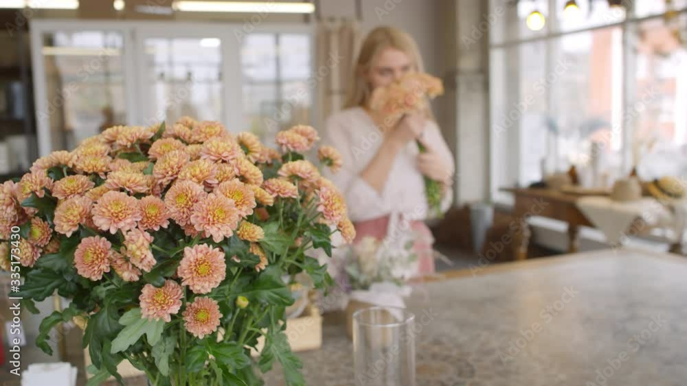 Handheld shot of female florist handing carnations to happy blond woman in flower shop. Bouquet of chrysanthemums standing in vase on counter Handheld shot of female florist handing carnations to happy blond woman in flower shop. Bouquet of chrysanthemums standing in vase on counter