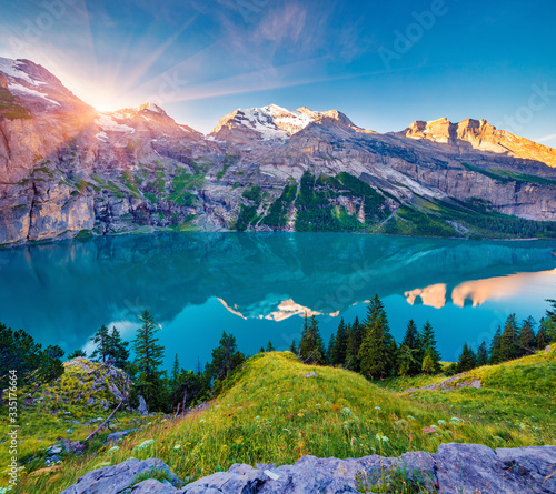 First sunlight gloving mountain hills and meadows. Picturesque summer view of unique Oeschinensee Lake. Sunrise in Swiss Alps with Bluemlisalp mountain on background, Switzerland, Europe.