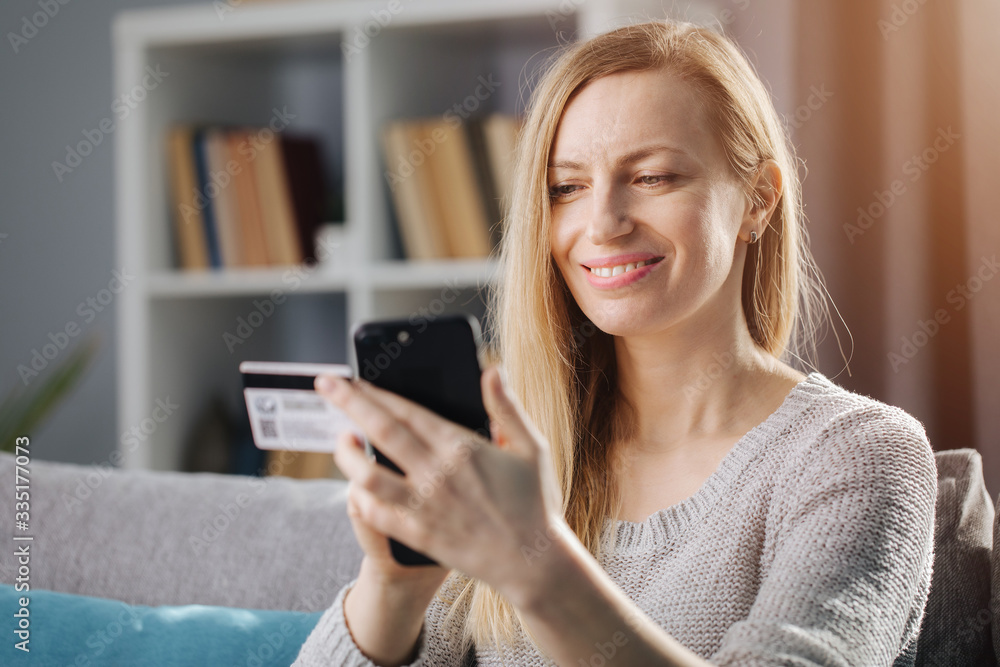 Attractive mature woman with long blond hair doing online shopping while sitting at home. Smiling lady in casual clothing using credit card and smartphone for payment. Attractive mature woman with long blond hair doing online shopping while sitting at home. Smiling lady in casual clothing using credit card and smartphone for payment.