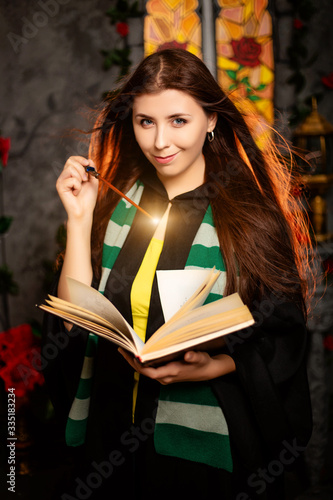 Fototapeta A young woman in a black mantle with a striped scarf around her neck casts a spell from a book and conjures with a wand of magic