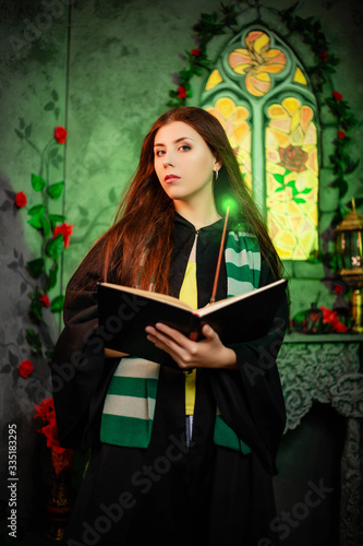 Fotografie, Obraz A young woman in a black mantle with a striped scarf around her neck casts a spell from a book and conjures with a wand of magic