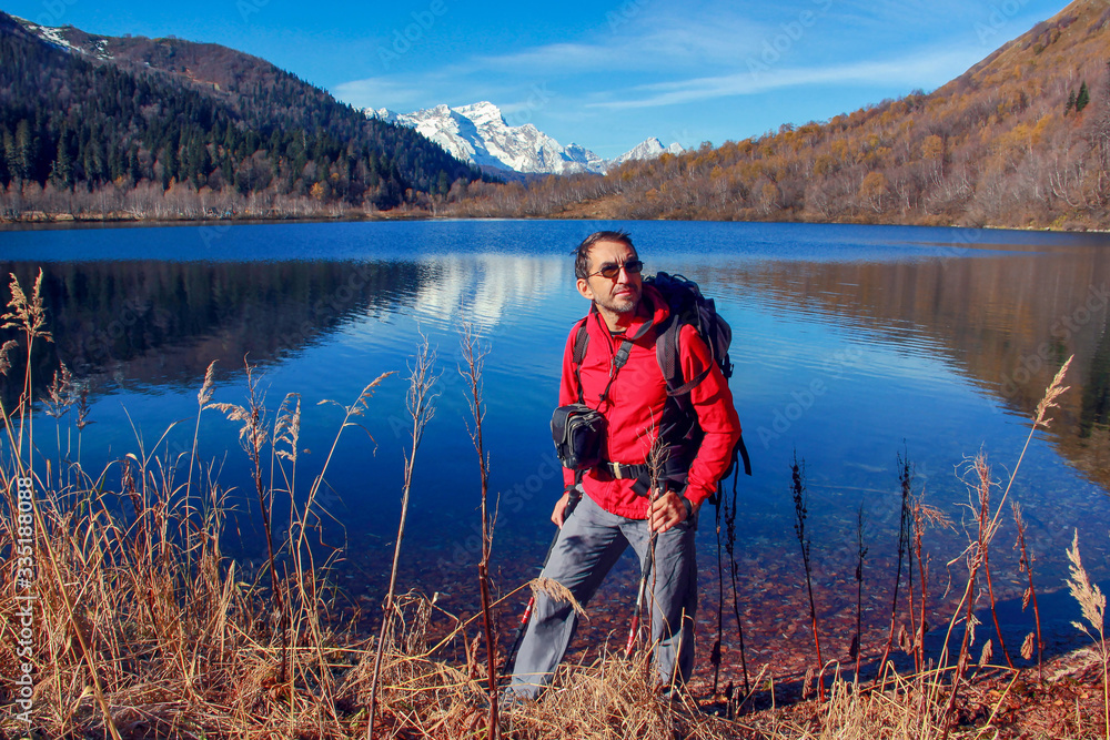 A middle-aged man in a red jacket and glasses with a backpack with trekking poles for tourism on the background of the lake which reflects the snow-capped mountains.